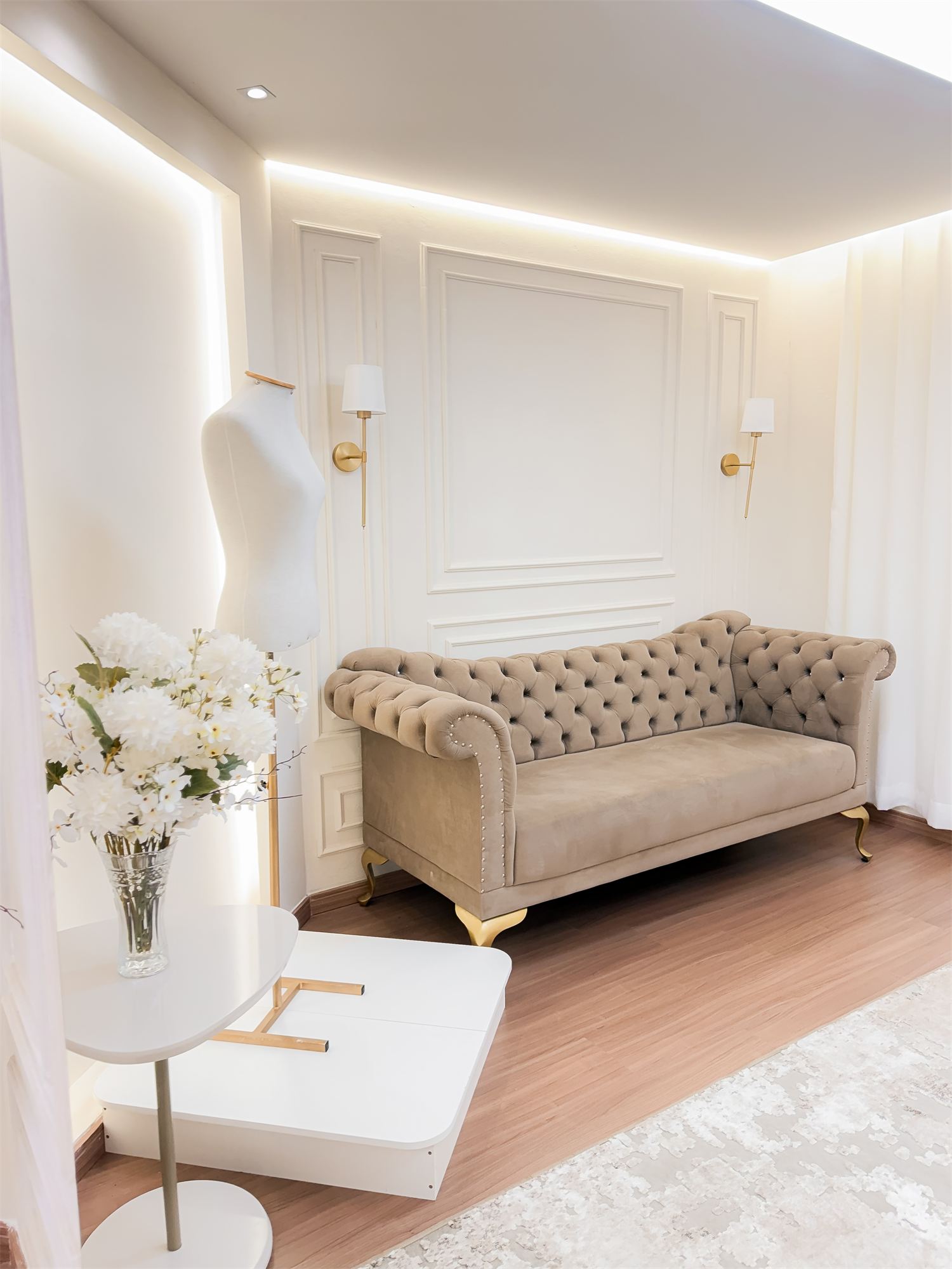 Sala de preparação com sofá capitonê, manequim, arandelas douradas e flores brancas