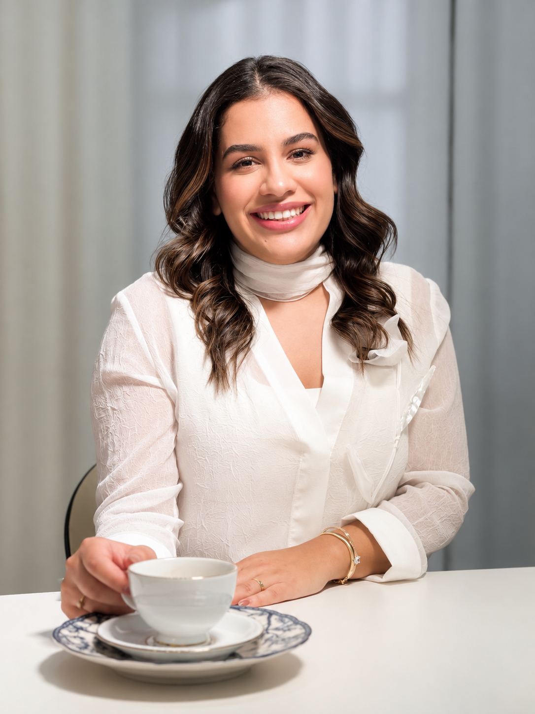Jacque Alvarenga sorrindo, de blusa branca, à mesa com uma xícara de porcelana
